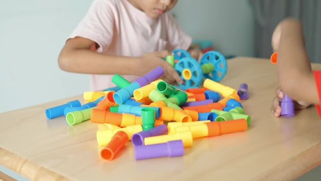 Adorable Asian boy, about 3-6 years old, wearing a pink shirt, happily playing with a puzzle shaped like a water pipe and having various toy items placed on a table inside his home, white background.