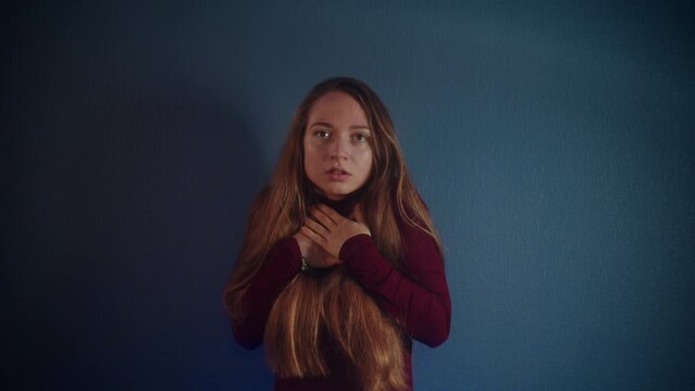 Portrait of a young scared trembling caucasian girl with long brown hair in a burgundy sweater looking at the camera and hides her mouth with her hands on a blue background. Emotion of fear and horror