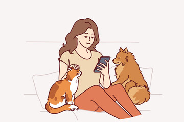 Woman owner of dog and cat sits on couch and plays on phone, enjoying spending time with pets. Girl strokes pets in need of affection and attention, to advertise shelter of domestic animals