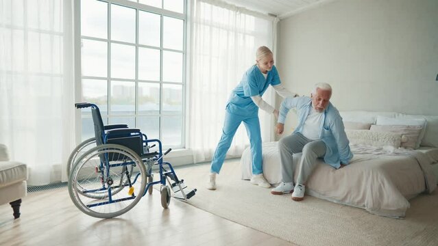 Nurse assisting senior man to get up bed.Assistant supporting patient while getting up bed and move towards wheelchair. Helping disabled man.Concept of comprehensive rehabilitation of disabled people