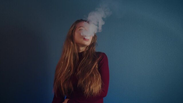 Portrait of a young beautiful caucasian blowing off steam girl with long brown hair in a burgundy sweater on a blue background in slow motion. The concept of vaping and smokong harm to health