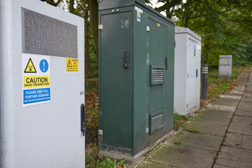 A row of telecommunications cabinets are lined up beside a pavement.  One of the cabinets in the foreground has stickers warning about radio transmissions and high electricity voltages
