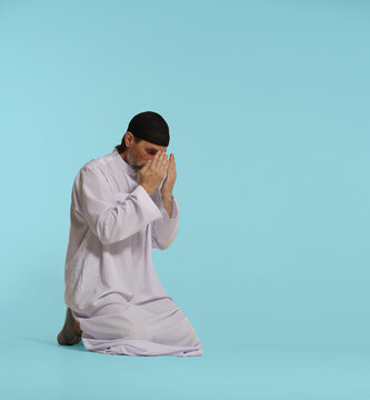 devout Muslim man kneels in pray, hands covering face in act of Namaz, fundamental Islamic ritual. Isolated against blue background. Faith, spirituality, and devotion in practice of Islamic prayer.