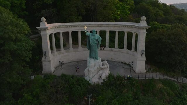 Pull back footage of historic tourist monument with St. Gellert statue in park. Revealing trees and paths. Cars driving on road around. Budapest, Hungary