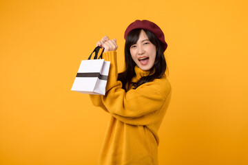 Young Asian woman in her 30s, holding a shopping paper bag, celebrating her shopping success in a fashionable yellow sweater and red beret against a yellow background.