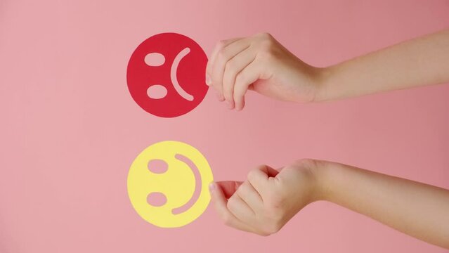 Vertical video of female hands holding happy and angry face paper, isolated on pink wall. feedback rating, Emotional intelligence, emotion control, mental health assessment, bipolar disorder concept