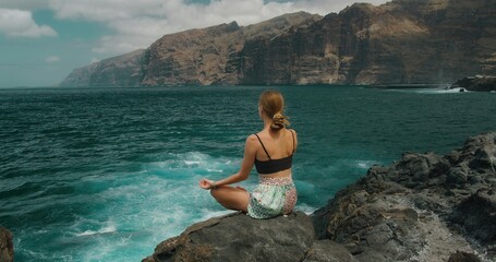 Woman meditates in lotus position on rocky shore by ocean. Girl breathing deeply sitting on...