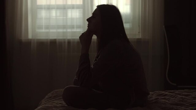 Silhouette of a millennial woman sitting on bed in the dark bedroom, depression, pain. Portrait of Unhappy sad woman sitting against window and thinking about her life