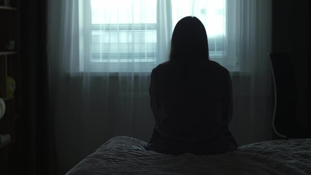 Back view Silhouette of a millennial woman sitting on bed in the dark bedroom, depression, pain. Portrait of Unhappy sad woman sitting against window and thinking about her life