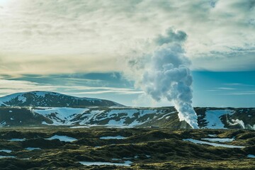 Geyser vent in Iceland surrounded by rocky snowy mountains covered with green grass