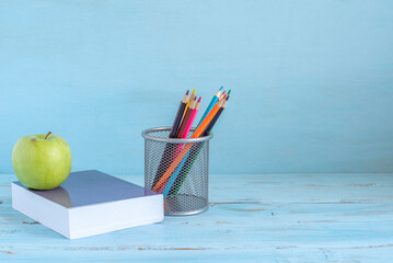 Green apple, study book, pencil holder with colorful pencils over blue wooden background with copy space. Back to school concept