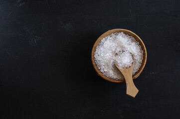 Coarse grainy salt in a wooden cup with a spoon on a black background, top view. Copy space, daylight