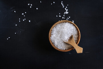 Coarse sea salt in a wooden cup with a spoon on a black background with a top view. Copy space, daylight