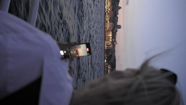 Young woman taking pictures of sea and coastal city while traveling on water by boat or ferry in evening or at night. Lights of city are burning. Using camera on your smartphone during boat cruise