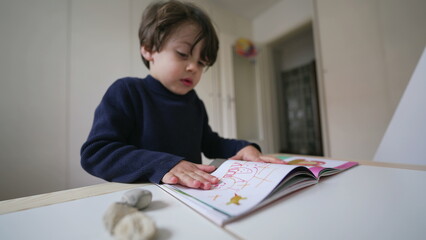 Child turning book pages looking at images and text. Little boy reading kindergarten school material