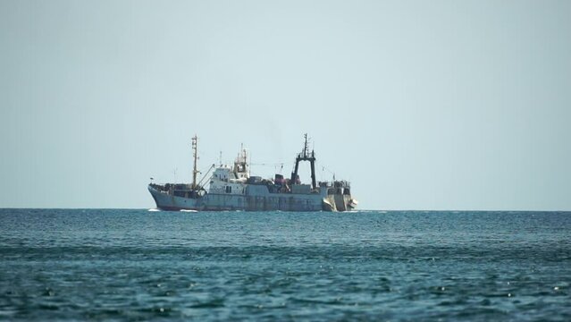 Fishing boat trawler catches fish while sailing on sea. A commercial fishing boat on the horizon in a distance sail to catch school of fish on calm sea surface in summer. Commercial catch of sea fish.