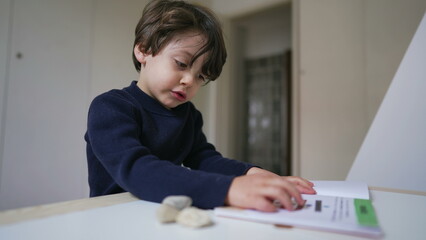 Small boy reading book sitting at desk in bedroom. Curious child studying by himself turning page and looking at text and images