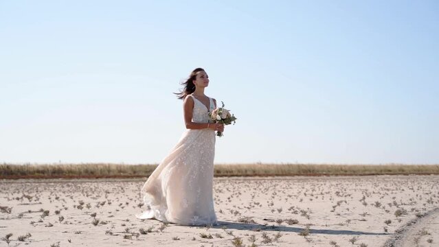 Pretty young bride holding delicate bouquet at desert landscape, windy weather. Elegance lady waiting for a groom after wedding ceremony. Attractive model posing in gorgeous white dress in nature.