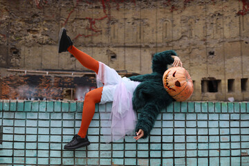 Halloween. a girl in a violet-blue dress, a green fur coat and orange tights and with a pumpkin instead of a head stands and sits on a blue brick wall. Halloween costume. ruins in the background - Powered by Adobe