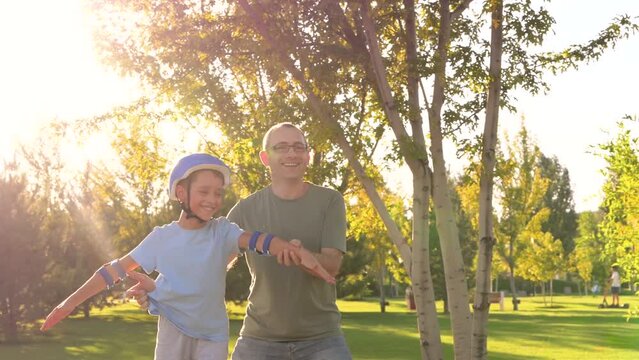 Happy father and child skateboarding in a green park. Boy in helmet learns to skateboard, dad coaches his son with his hands helping him. Father and son. Active recreation urban environment