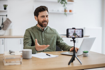 Confident bearded man sitting at desk and working on wireless laptop while recording video on modern smartphone fixed on tripod. Male freelancer creating new content for social media at home. - obrazy, fototapety, plakaty