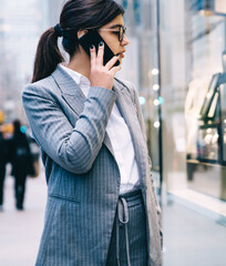 Fototapeta premium Confident formal young beautiful business lady talking on smartphone while walking on street