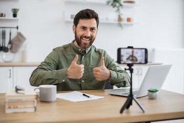 Confident bearded man sitting at desk and working on wireless laptop while recording video on modern smartphone fixed on tripod showing thumbs up