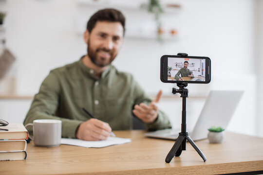 Blurred background of smiling man taking notes while communicating with followers in social media during live streaming at home. Popular blogger using laptop, smartphone and tripod for online work.