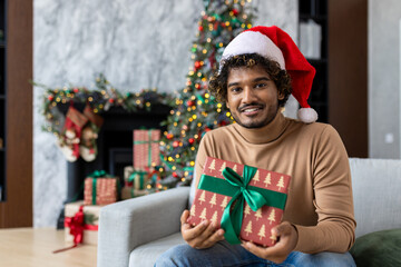 Online video call, holiday greeting for Christmas, man smiling and looking at camera in red Santa hat, sitting on sofa in living room, showing gift, Hispanic man greeting friends with New Year.