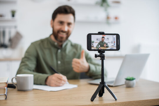 Blurred background of smiling man taking notes while communicating with followers in social media during live streaming at home showing thumb up