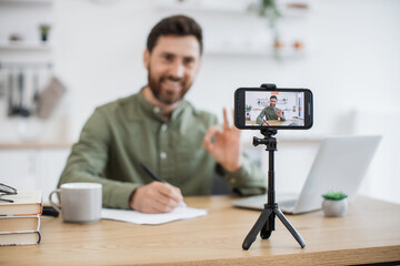 Blurred background of smiling man taking notes while communicating with followers in social media during live streaming at home showing sign ok