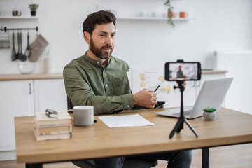 Economic male expert presenting graphic calculation while sitting at home office and looking on mobile camera. Handsome man creating new video for business vlog with help of smartphone and tripod.