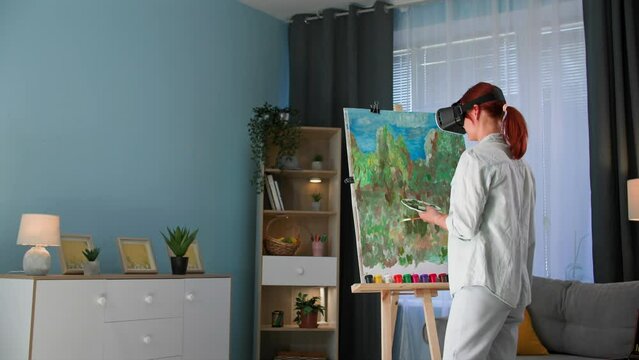 virtual art, talented female artist in viar glasses draws a picture on canvas using paints and a brush while standing in a cozy room