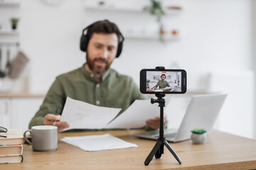 Focused male blogger in wireless headphones examining financial graphs and explaining to followers all peculiarities. Caucasian man using modern smartphone and tripod for recording video at home.