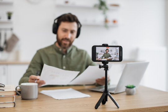 Focused male blogger in wireless headphones examining financial graphs and explaining to followers all peculiarities. Caucasian man using modern smartphone and tripod for recording video at home.