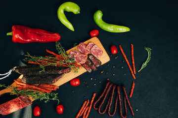 Sausages of different types and sizes on a wooden tray with green paper and tomatoes. Set of smoked sausage on dark background. Top view