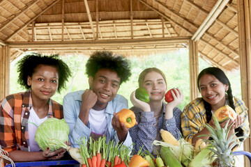 Group of happy Multiethnic teenager friend holding organic food products from their own farm at local market fair, smiling young diverse farmer selling fresh harvest fruit and vegetable at marketplace
