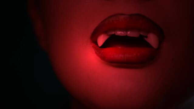 Sexy Vampire Woman's red bloody lips close-up. Vampire girl licking fangs with tongue. Fashion Glamour Halloween art design. Close up of female vampire mouth, teeth. On black background. Strobe lights