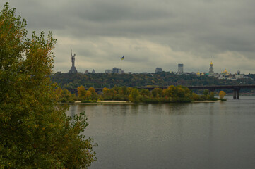 Autumn landscape of Dnipro River in Kyiv during cloudy day. Famous Kyiv's hills on right bank of river.Kyiv Pechersk Lavra, residential buildings and Motherland Monument on horizon.Tree leaves border