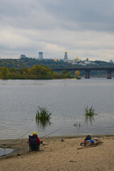 Fishing in comfort. Man sits in a folding chair and fishes on the bank of the Dnipro river in Kyiv. Cloudy autumn day. Famous Kyiv's hills with Kyiv Pechersk Lavra on right bank of river on horizon