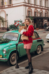 Young beautiful blonde woman in a red knitted cardigan and warm mittens holds a Christmas bouquet while walking through an old European city