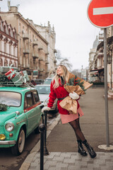 A young blonde woman walks down the street with a Christmas bouquet near a retro car decorated with Christmas boxes