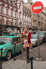 A young blonde woman walks down the street with a Christmas bouquet near a retro car decorated with Christmas boxes