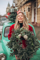 Beautiful young blonde woman in a red knitted cardigan holding a Christmas wreath while sitting on the hood of a retro car