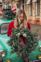 Beautiful young blonde woman in a red knitted cardigan holding a Christmas wreath while sitting on the hood of a retro car