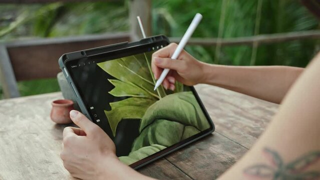 Female artist hands drawing with stylus on tablet at the cafe on tropical island, palm trees. Young designer works remotely open air at exotic location. Pretty woman editing illustration. Close-up.