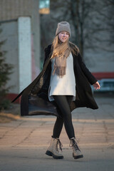 Fototapeta premium Portrait of a young beautiful blonde girl in a knitted hat and dark coat in an urban environment.