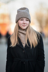 Portrait of a young beautiful blonde girl in a knitted hat and dark coat in an urban environment.