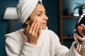 Hispanic woman with white towel on her head washes face, cleans skin with soap or soft foam....