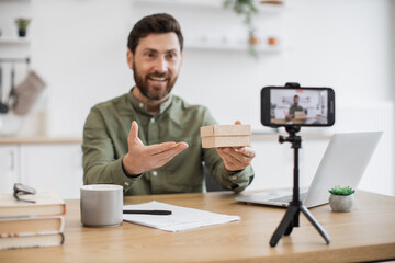 Positive content maker sitting at home office and showing on mobile camera small cardboard box during live streaming. Caucasian man sharing with subscribers unpacking process of new purchase.